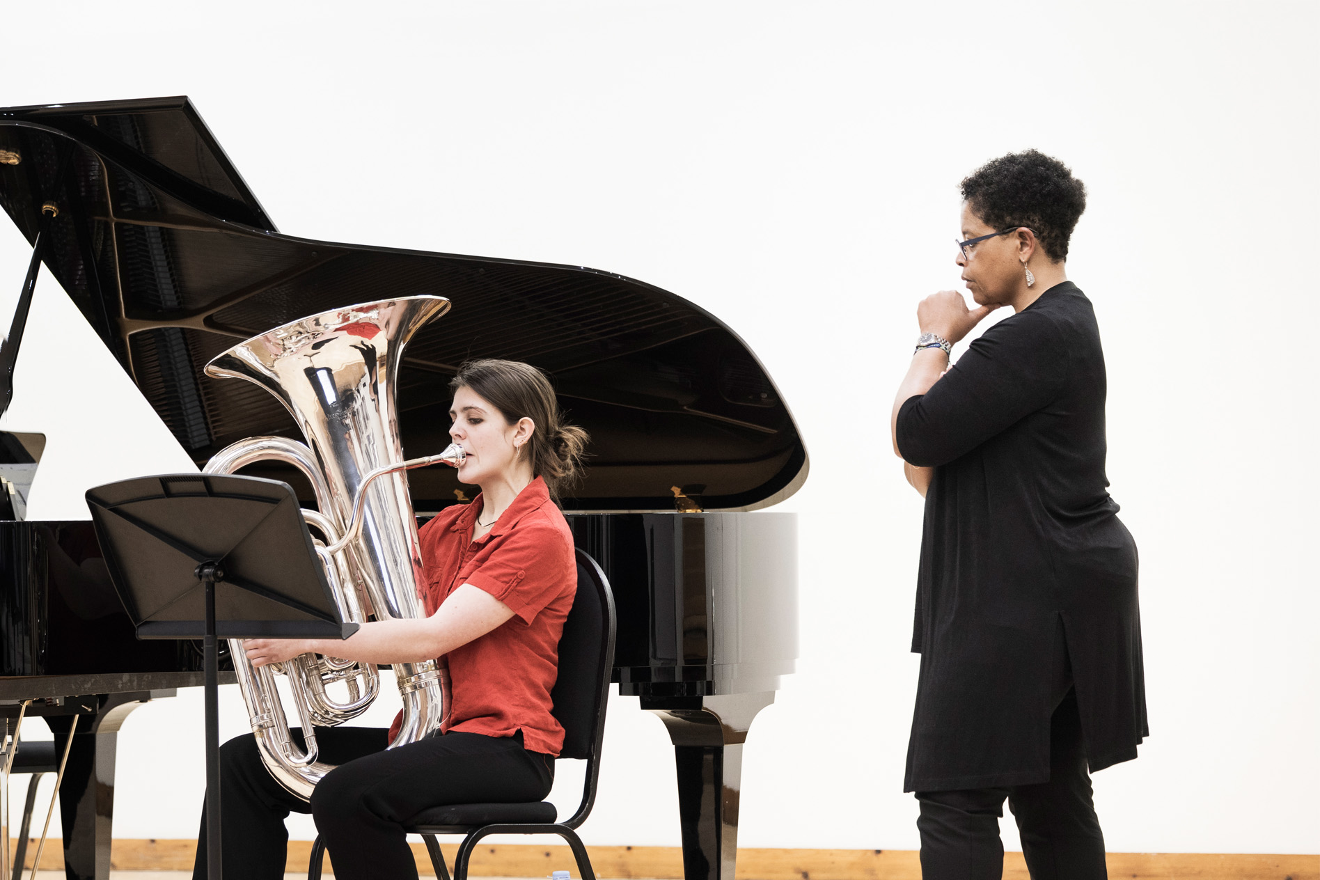 Female student playing her tuba, while a women with a black cardigan observing. A piano is in the background to accompany her performance.